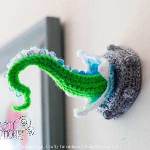 May include: Crocheted wall art featuring a gray, textured creature with a green tentacle emerging. The tentacle is adorned with blue and white beads. The creature's mouth is white and blue. A PDF icon is in the lower right corner.
