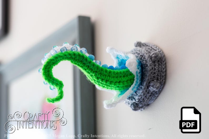 May include: Crocheted wall art featuring a gray, textured creature with a green tentacle emerging. The tentacle is adorned with blue and white beads. The creature's mouth is white and blue. A PDF icon is in the lower right corner.