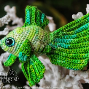 Puede incluir: Pez betta amigurumi de crochet verde con un degradado de color verde claro y azul. El pez tiene ojos grandes y negros con un iris azul. El pez est&aacute; sentado sobre una superficie blanca similar al coral.
