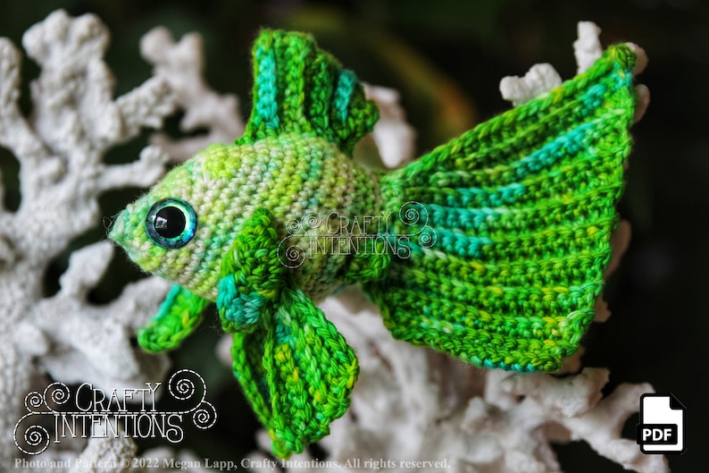 Puede incluir: Pez betta amigurumi de crochet verde con un degradado de color verde claro y azul. El pez tiene ojos grandes y negros con un iris azul. El pez est&aacute; sentado sobre una superficie blanca similar al coral.
