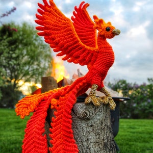 May include: A crocheted phoenix with orange and red feathers, perched on a wooden stump. The phoenix has a long tail and its wings are spread out. The background is a blurry image of a green grassy field and a cloudy sky.
