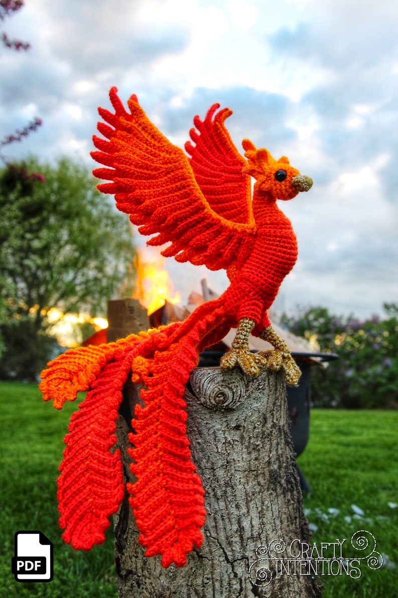 May include: A crocheted phoenix with orange and red feathers, perched on a wooden stump. The phoenix has a long tail and its wings are spread out. The background is a blurry image of a green grassy field and a cloudy sky.