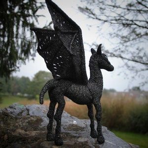 May include: Black crocheted winged horse sculpture standing on a rock. The sculpture is made of black yarn and has a detailed design. The sculpture is a great addition to any home decor.