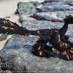 May include: A black crochet dinosaur with rainbow accents. The dinosaur is standing on a rock with a sandy beach in the background. The image includes the text "CRAFTY INTENTIONS".