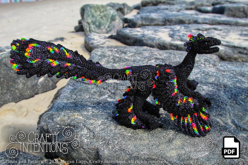 May include: A black crochet dinosaur with rainbow accents. The dinosaur is standing on a rock with a sandy beach in the background. The image includes the text "CRAFTY INTENTIONS".