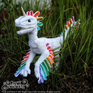 May include: A white crocheted unicorn dinosaur with a rainbow mane and tail. The dinosaur is standing in a bed of green grass.