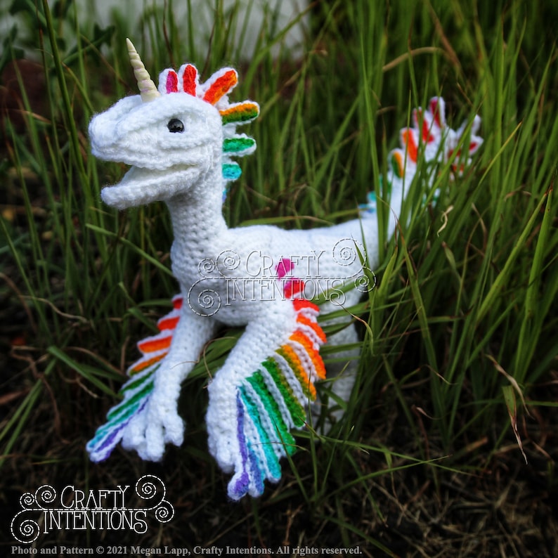 May include: A white crocheted unicorn dinosaur with a rainbow mane and tail. The dinosaur is standing in a bed of green grass.