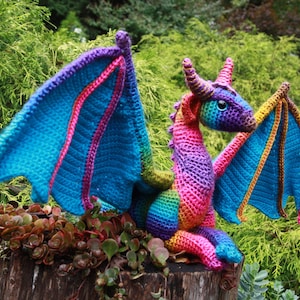 K&ouml;nnte beinhalten: Ein bunter H&auml;kel-Drache mit regenbogenfarbenen Schuppen und blauen Fl&uuml;geln. Der Drache sitzt auf einem Baumstumpf mit gr&uuml;nen Pflanzen im Hintergrund.