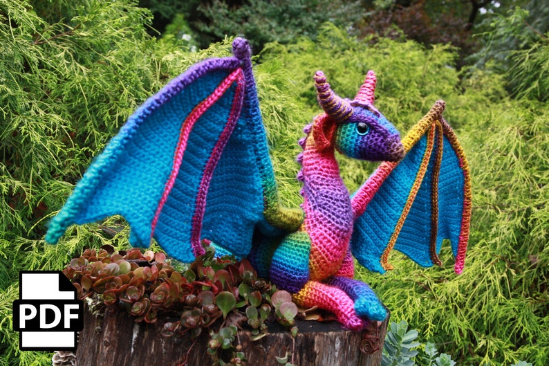 K&ouml;nnte beinhalten: Ein bunter H&auml;kel-Drache mit regenbogenfarbenen Schuppen und blauen Fl&uuml;geln. Der Drache sitzt auf einem Baumstumpf mit gr&uuml;nen Pflanzen im Hintergrund.
