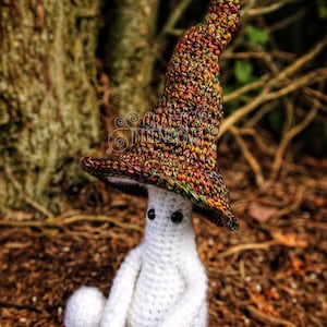 May include: A white crocheted ghost wearing a colorful crocheted witch hat. The ghost is sitting on a bed of brown leaves.