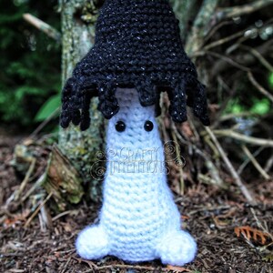 May include: A crocheted mushroom amigurumi toy with a black cap and white body. The mushroom has googly eyes and is sitting on a bed of pine needles.