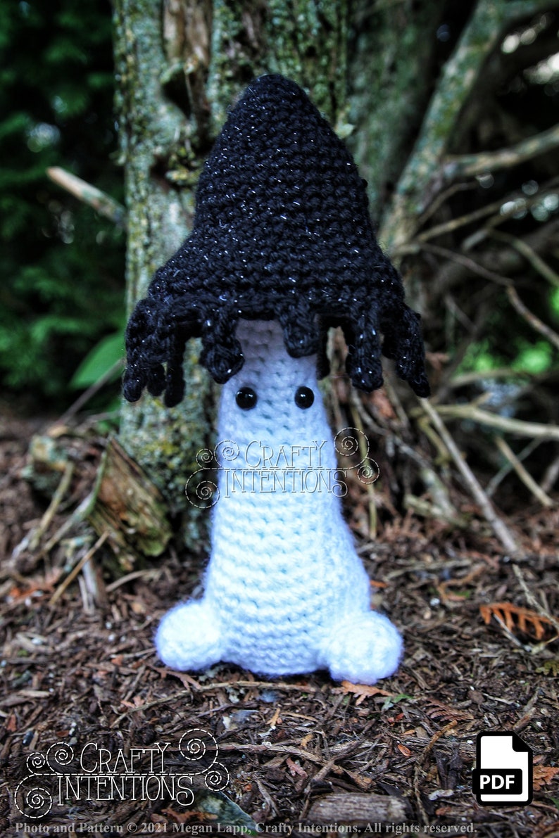 May include: A crocheted mushroom amigurumi toy with a black cap and white body. The mushroom has googly eyes and is sitting on a bed of pine needles.