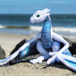 May include: A crocheted white and blue dragon sitting on a rock with the ocean in the background. The dragon has a white body with blue wings and a blue tail. The dragon has green eyes and a white snout. The dragon is sitting on a rock with the ocean in the background. The ocean is blue and there are waves crashing on the shore. The dragon is looking to the right. The image includes the text "CRAFTY INTENTIONS" and a PDF icon.