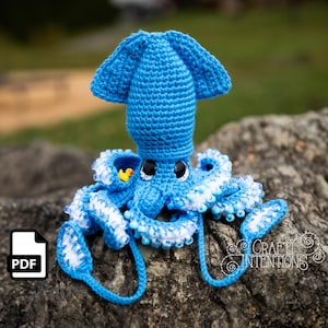 May include: A crocheted blue squid with white and blue accents. The squid has large black eyes and a small yellow duck toy in its tentacles. The squid is sitting on a rock.