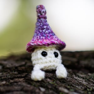 Puede incluir: Una figura de hongo de ganchillo hecha a mano con cuerpo blanco, ojos negros y gorro morado y rosa. El hongo tiene peque&ntilde;as patas blancas y est&aacute; sobre un fondo natural borroso. La imagen incluye el texto "Crafty Intentions" y un icono PDF.