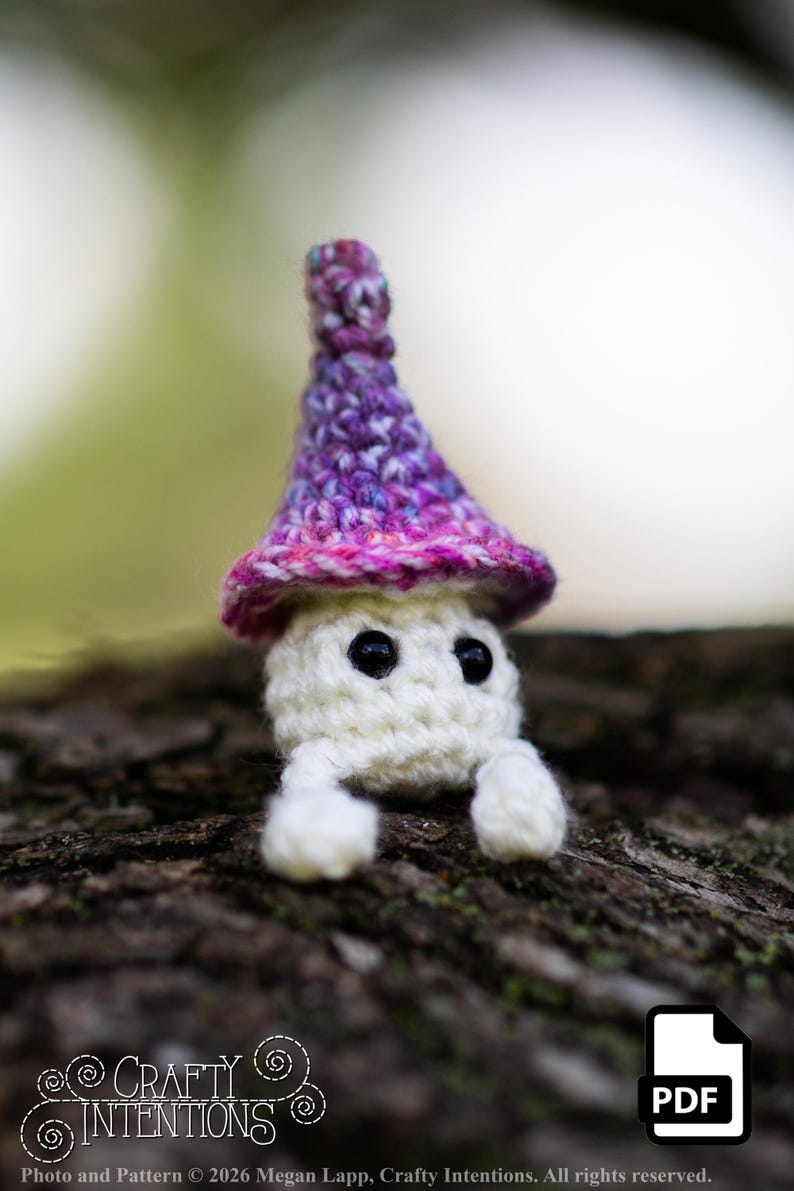 Puede incluir: Una figura de hongo de ganchillo hecha a mano con cuerpo blanco, ojos negros y gorro morado y rosa. El hongo tiene peque&ntilde;as patas blancas y est&aacute; sobre un fondo natural borroso. La imagen incluye el texto "Crafty Intentions" y un icono PDF.