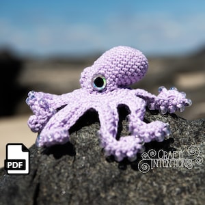 Puede incluir: Un pulpo de lavanda tejido a ganchillo con grandes ojos iridiscentes y puntas de tentáculos con cuentas. El pulpo está sentado sobre una roca gris oscura. La imagen incluye un icono PDF y el texto "Crafty Intentions".