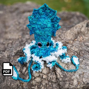 May include: A handmade crochet octopus in shades of blue and white. The octopus has large, expressive eyes and a ruffled head. The tentacles are detailed with white edging. The image includes a PDF icon in the lower left corner and the text "Crafty Intentions" in the lower right.