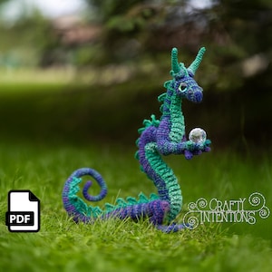 May include: A crocheted dragon figurine in shades of blue, green, and purple, holding a clear crystal ball. The dragon is posed on green grass. The image includes a PDF icon and the text "Crafty Intentions".