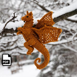 Puede incluir: Un dragón tejido a ganchillo en tonos naranja, con alas detalladas y una cola enroscada, posado en una rama cubierta de nieve. El dragón tiene un aspecto texturizado y se encuentra sobre un fondo invernal borroso. Un icono PDF está en la esquina inferior izquierda.