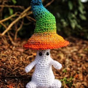May include: A white crocheted ghost wearing a rainbow crocheted hat. The hat has a pointed top and a wide brim. The ghost is sitting on a bed of brown pine needles.