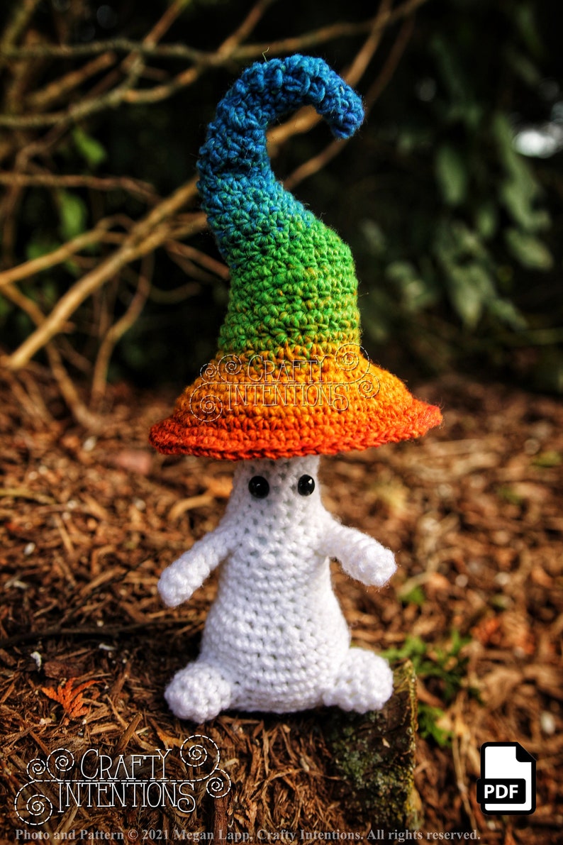 May include: A white crocheted ghost wearing a rainbow crocheted hat. The hat has a pointed top and a wide brim. The ghost is sitting on a bed of brown pine needles.
