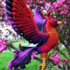 May include: A crocheted phoenix bird with red, orange, purple, and black yarn. The bird is perched on a branch with pink blossoms.