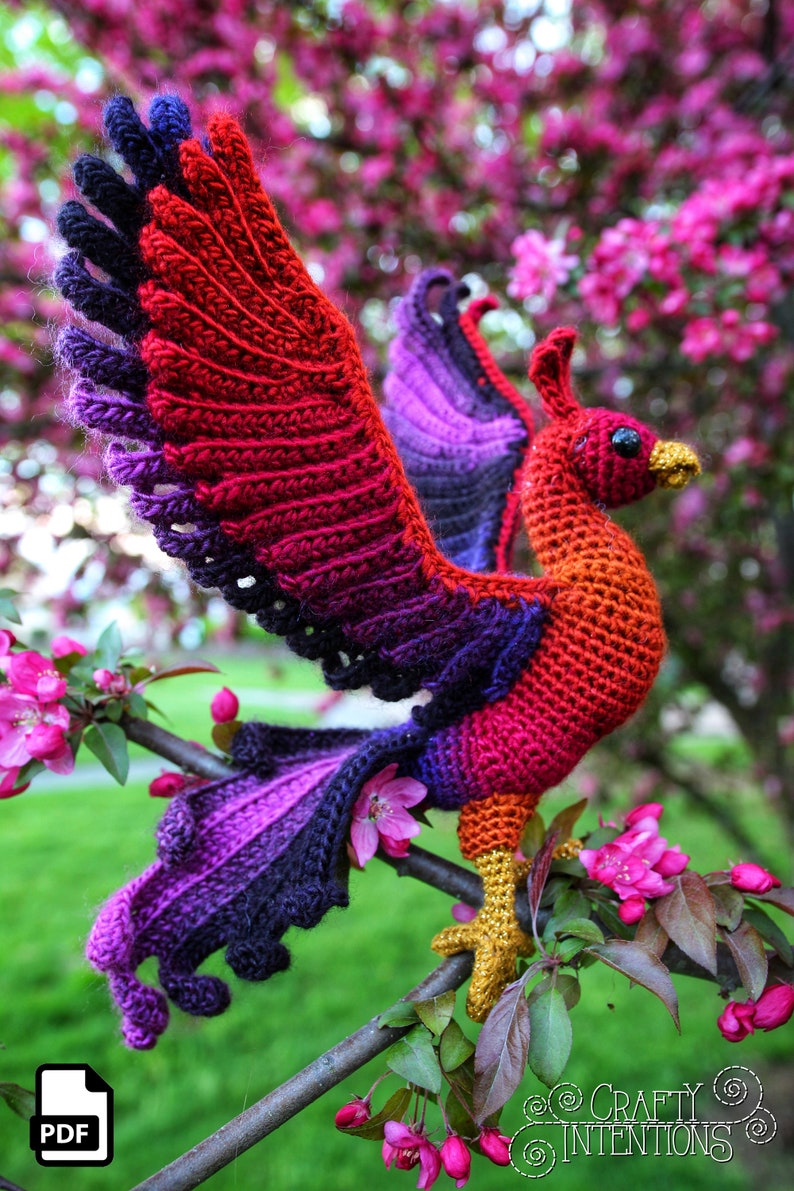 May include: A crocheted phoenix bird with red, orange, purple, and black yarn. The bird is perched on a branch with pink blossoms.