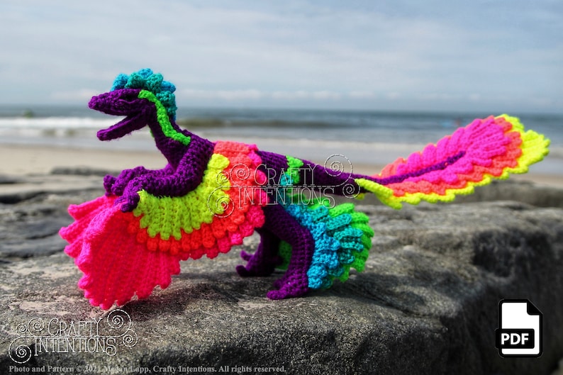May include: A crocheted dinosaur toy with a rainbow colour scheme. The dinosaur is posed with its wings spread out. The toy is sitting on a rock with a beach and ocean in the background.