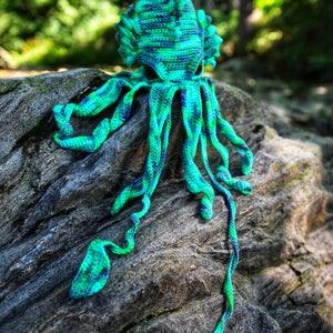 May include: Crocheted octopus amigurumi toy with a green and blue color scheme. The octopus is sitting on a rock with a green and brown color scheme. The octopus has a long, thin tentacle that is hanging down. The image includes the text "CRAFTY INTENTIONS" and a PDF icon.