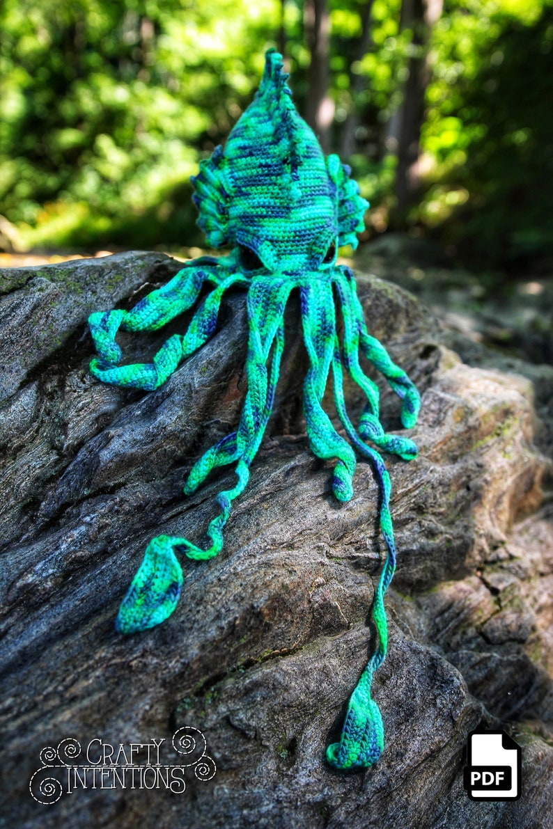 May include: Crocheted octopus amigurumi toy with a green and blue color scheme. The octopus is sitting on a rock with a green and brown color scheme. The octopus has a long, thin tentacle that is hanging down. The image includes the text "CRAFTY INTENTIONS" and a PDF icon.
