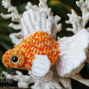 Puede incluir: Juguete amigurumi de ganchillo en forma de pez dorado, hecho con hilo naranja y blanco. El juguete tiene un fondo blanco y est&aacute; sentado sobre una superficie blanca similar al coral. El juguete tiene un ojo de bot&oacute;n negro.