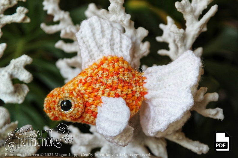 Puede incluir: Juguete amigurumi de ganchillo en forma de pez dorado, hecho con hilo naranja y blanco. El juguete tiene un fondo blanco y est&aacute; sentado sobre una superficie blanca similar al coral. El juguete tiene un ojo de bot&oacute;n negro.