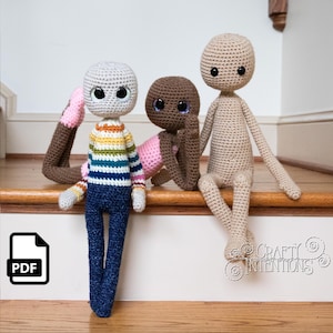 May include: Three crocheted dolls sitting on a wooden staircase. The dolls are different colours and have different outfits. The doll on the left is wearing a striped shirt and blue jeans. The doll in the middle is wearing a pink dress. The doll on the right is wearing no clothes. The dolls are all smiling and have big eyes.