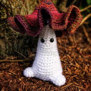 May include: A crocheted mushroom amigurumi toy with a white body and a red, purple, and green cap. The toy has black eyes and is sitting on a bed of brown mulch.