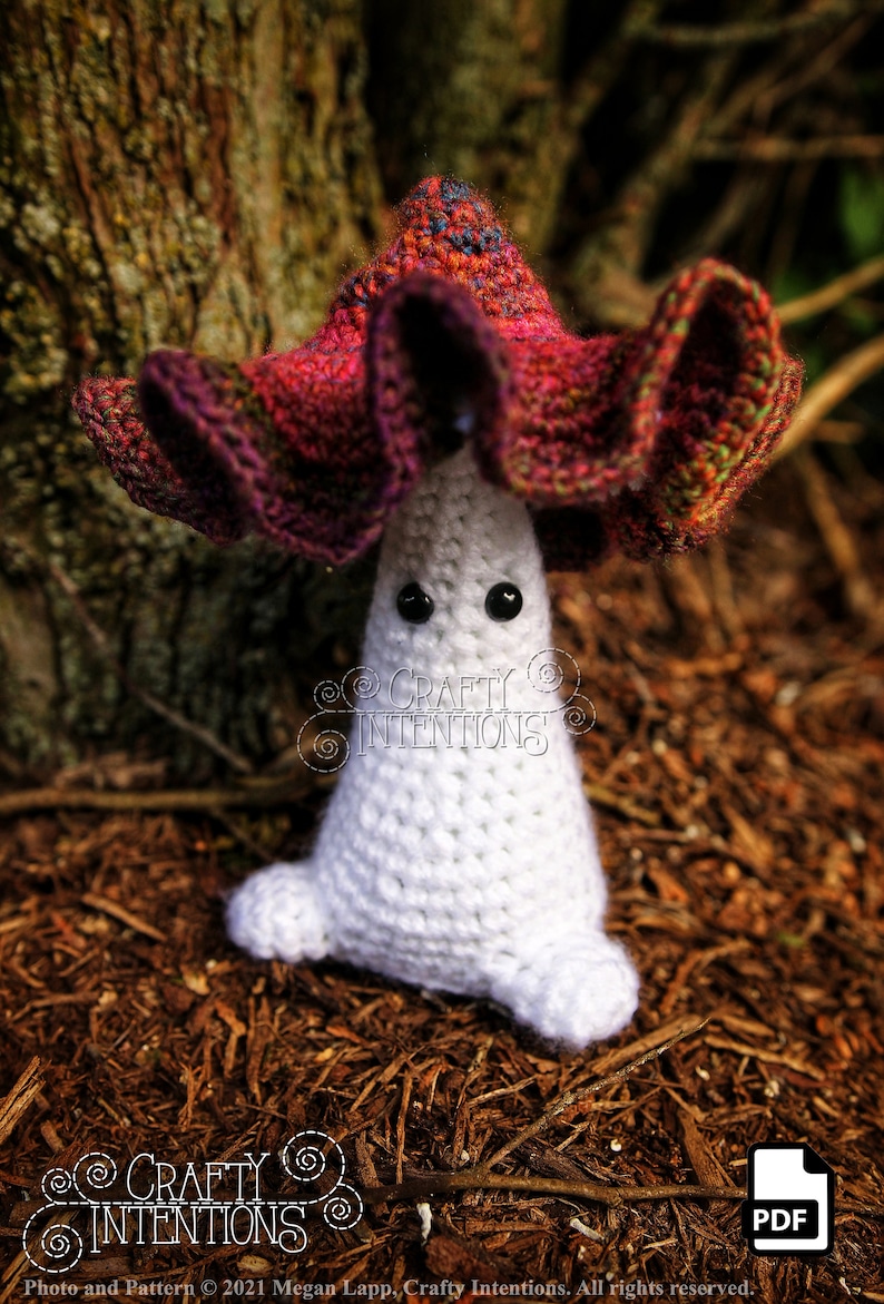 May include: A crocheted mushroom amigurumi toy with a white body and a red, purple, and green cap. The toy has black eyes and is sitting on a bed of brown mulch.