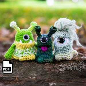 Peut inclure: Trois monstres amigurumi en crochet avec de grands yeux. Les monstres sont verts, bleus et gris avec des traits du visage différents. Les monstres sont assis sur une surface en bois. L'image comprend le texte "CRAFTY INTENTIONS".