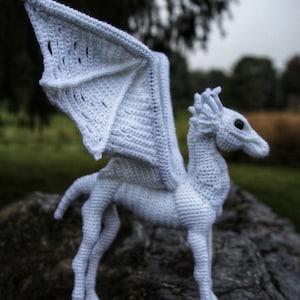 May include: White crocheted dragon with wings outstretched, standing on a tree stump. The dragon has a long neck and a detailed head with a pointed snout.