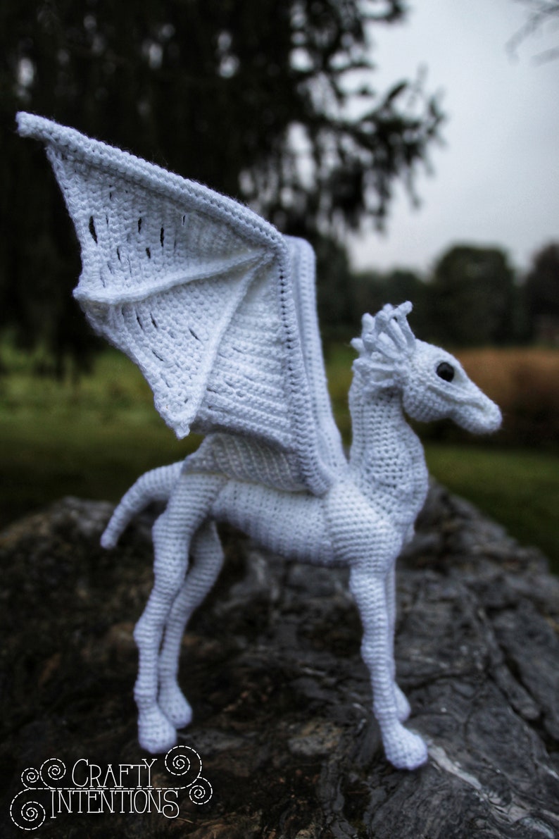 May include: White crocheted dragon with wings outstretched, standing on a tree stump. The dragon has a long neck and a detailed head with a pointed snout.