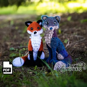May include: Two crocheted stuffed animals, a fox and a wolf, sitting on a bed of green grass. The fox is orange, white, and black, while the wolf is blue, purple, and grey. The fox has a white tip on its tail. The wolf has a blue and purple tail. The image includes the text "Crafty Intentions".