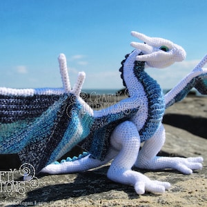May include: A white crocheted dragon with blue and white wings. The dragon is sitting on a rock with the ocean in the background. The text "CRAFTY INTENTIONS" is visible in the image.