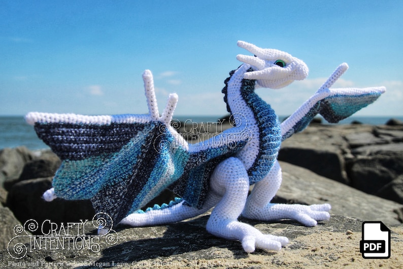 May include: A white crocheted dragon with blue and white wings. The dragon is sitting on a rock with the ocean in the background. The text "CRAFTY INTENTIONS" is visible in the image.