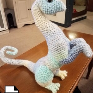 May include: A crocheted amigurumi dragon toy in shades of blue, white, and green. The dragon has a long neck, a curled tail, and a single black eye.
