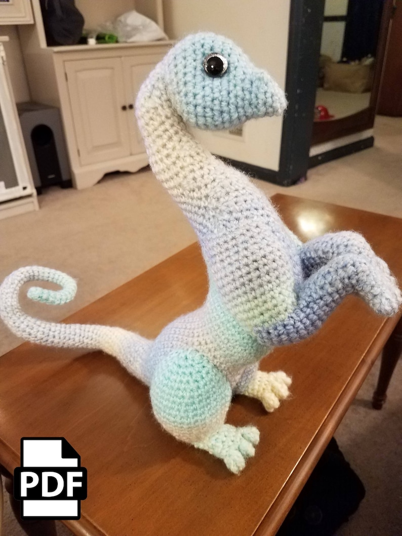 May include: A crocheted amigurumi dragon toy in shades of blue, white, and green. The dragon has a long neck, a curled tail, and a single black eye.