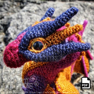 May include: Close-up of a crocheted dragon head with a purple, orange, and pink color scheme. The dragon has a black eye and a textured, sparkly finish.