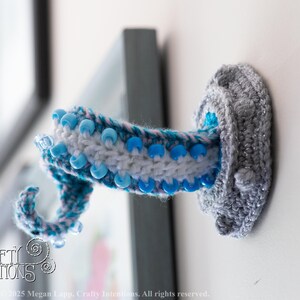 May include: A handmade, crochet octopus tentacle wall hook in shades of gray, white, and blue. The tentacle is adorned with blue beads and is mounted on a white wall. The hook is a unique, decorative item.