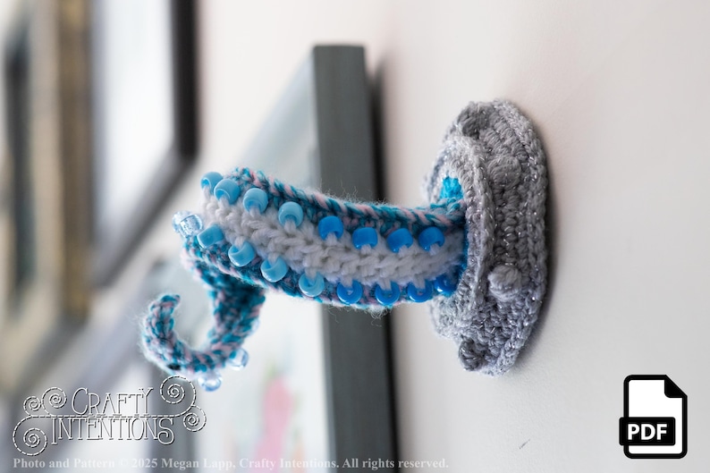 May include: A handmade, crochet octopus tentacle wall hook in shades of gray, white, and blue. The tentacle is adorned with blue beads and is mounted on a white wall. The hook is a unique, decorative item.