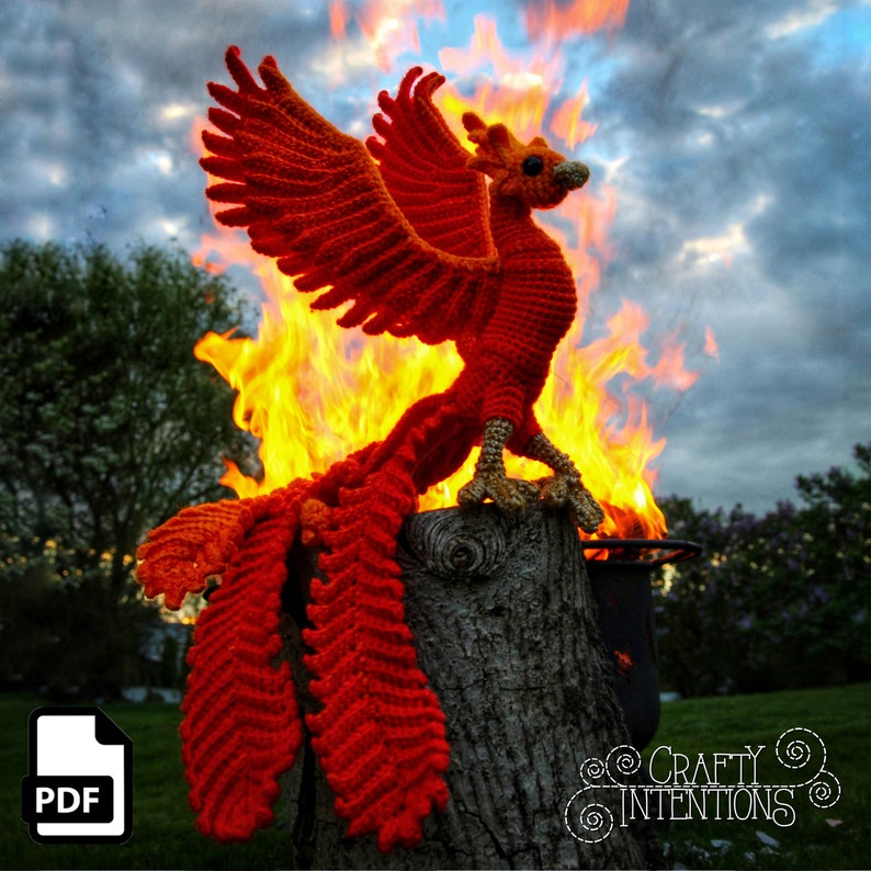 May include: A crocheted phoenix figurine in orange and red, perched on a tree stump with flames behind it. The phoenix has a long tail and wings, and its head is turned to the side. The image includes the text "Crafty Intentions".