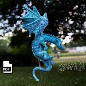 Op de afbeelding: Een gehaakt drakenfiguur met een kleurverloop van blauw en turquoise garen. Het wezen heeft grote, gedetailleerde vleugels, een lange gebogen staart en een getextureerd lichaam. De tekst Crafty Intentions is zichtbaar, samen met een PDF-pictogram.