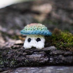 Puede incluir: Un hongo de ganchillo hecho a mano con un tallo blanco y un sombrero multicolor en tonos de azul, verde y rosa. El hongo tiene dos ojos negros y se encuentra sobre un fondo natural borroso. La imagen incluye el texto "Crafty Intentions" y un icono PDF.